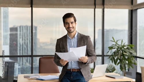 Happy young Latin business man checking financial documents in office. Smiling male professional account manager executive lawyer holding corporate tax bill papers standing at work photo