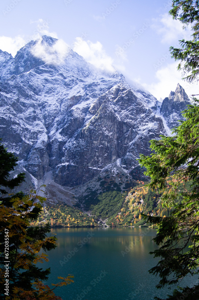 Serene view of Morskie Oko in Poland. The emerald lake reflects snow-capped Tatra peaks, bordered by vibrant autumn trees. A testament to nature's harmony and seasonal beauty.