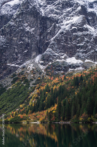 Fototapeta Naklejka Na Ścianę i Meble -  Stunning view of Morskie Oko in Poland. Snow-kissed cliffs rise over a tranquil lake, reflecting a forest ablaze with autumn colors. A perfect blend of winter and fall in nature's canvas.