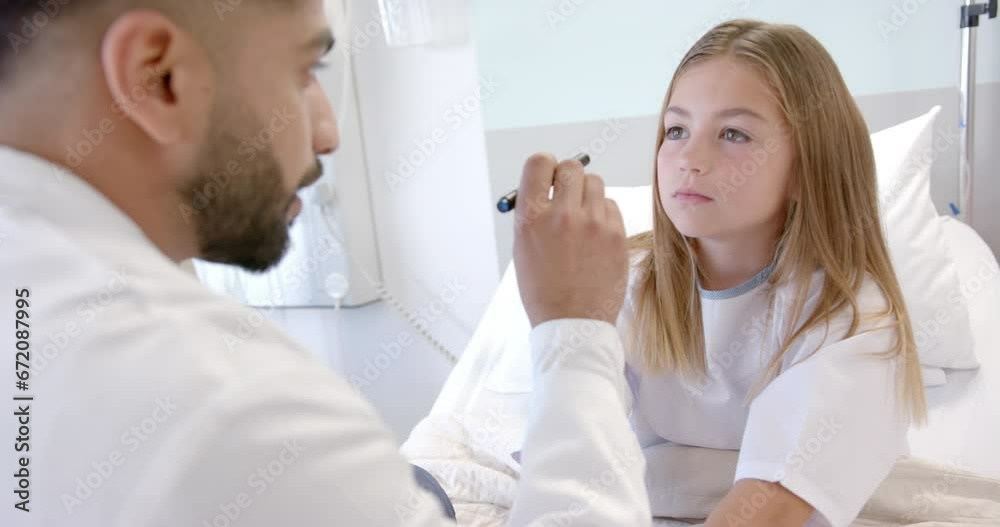 Diverse male doctor checking eyes of girl patient in hospital bed with penlight, slow motion Diverse male doctor checking eyes of girl patient in hospital bed with penlight, slow motion