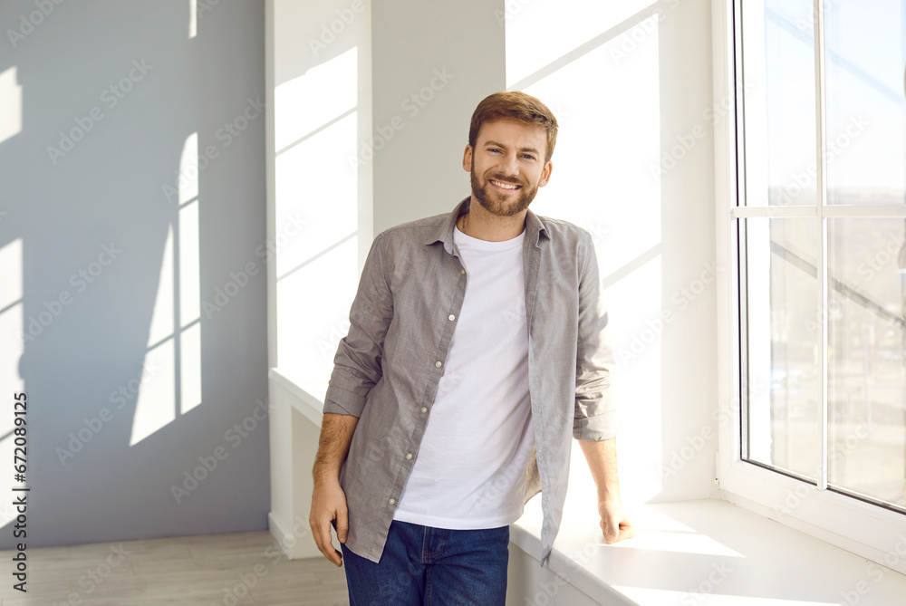 Portrait of a happy man at home. Smiling young man in a casual gray shirt, white T shirt and jeans standing by the window in a light clean room in his house or apartment Portrait of a happy man at home. Smiling young man in a casual gray shirt, white T shirt and jeans standing by the window in a light clean room in his house or apartment