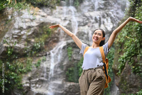Overjoyed female tourist with backpack raising arms in front of beautiful tropical waterfall