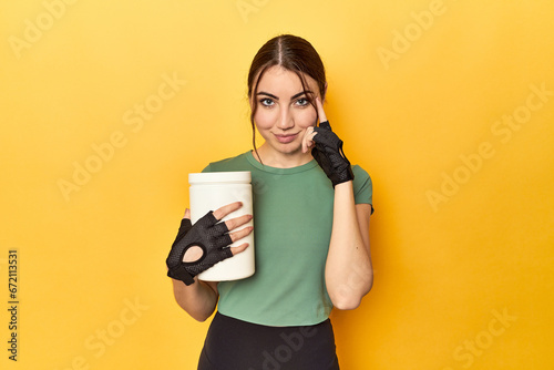 Fit woman holding a protein shake pointing temple with finger, thinking, focused on a task.
