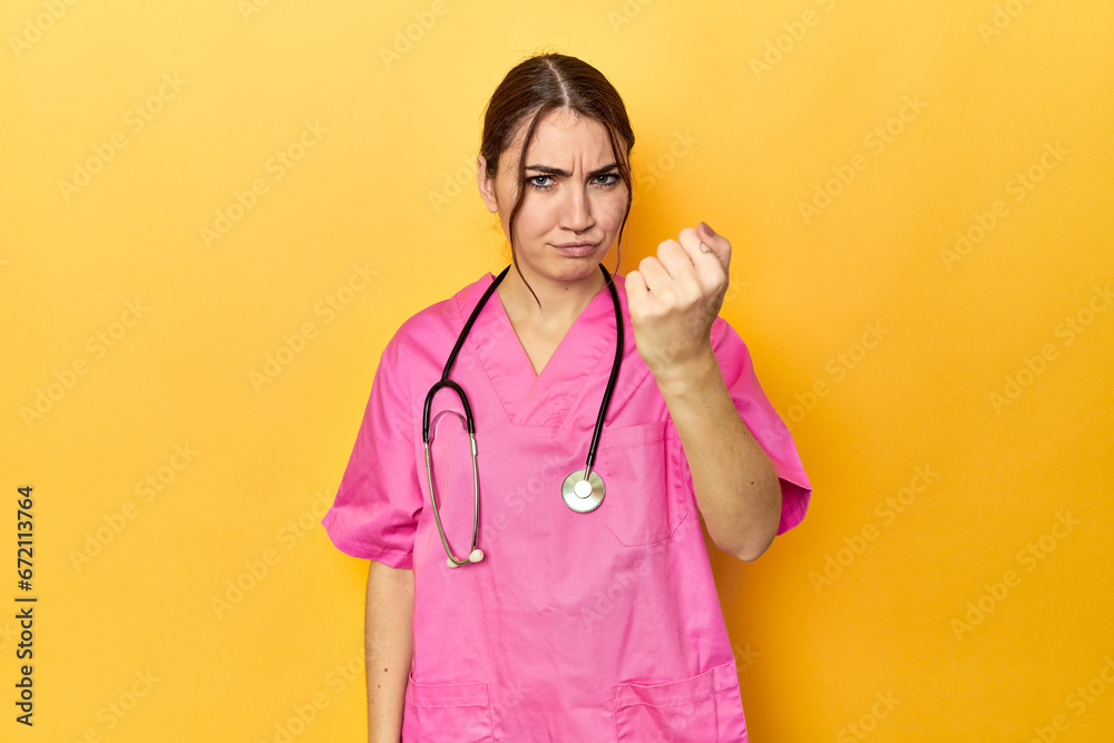 Nursing assistant in yellow background showing fist to camera, aggressive facial expression. Nursing assistant in yellow background showing fist to camera, aggressive facial expression.