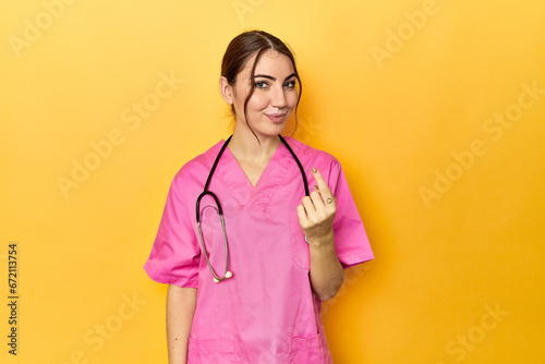 Nursing assistant in yellow background pointing with finger at you as if inviting come closer.