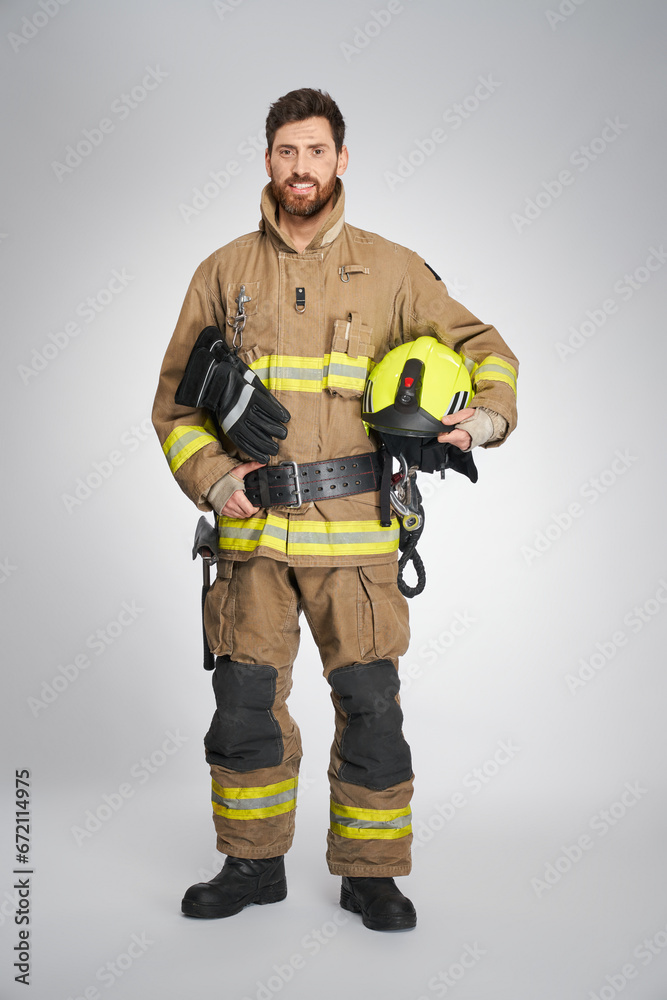 Strong, bearded fireman posing to camera with yellow protective helmet. Front view of happy male firefighter keeping hand on leather belt, while looking at camera, on gray background. Concept of work. Strong, bearded fireman posing to camera with yellow protective helmet. Front view of happy male firefighter keeping hand on leather belt, while looking at camera, on gray background. Concept of work.
