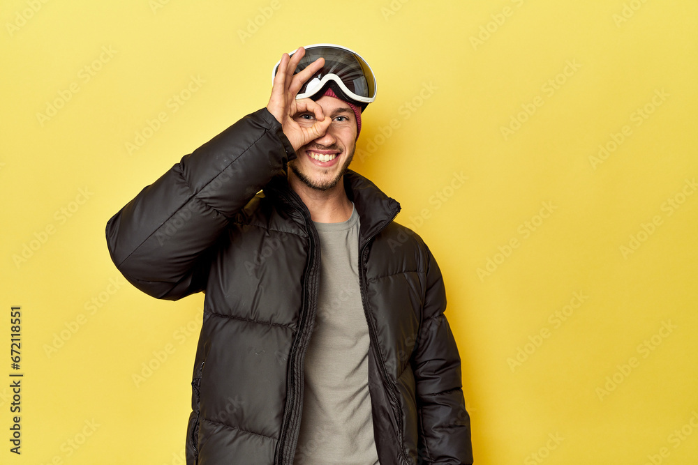 Man in winter coat with snow goggles on yellow excited keeping ok gesture on eye. Man in winter coat with snow goggles on yellow excited keeping ok gesture on eye.