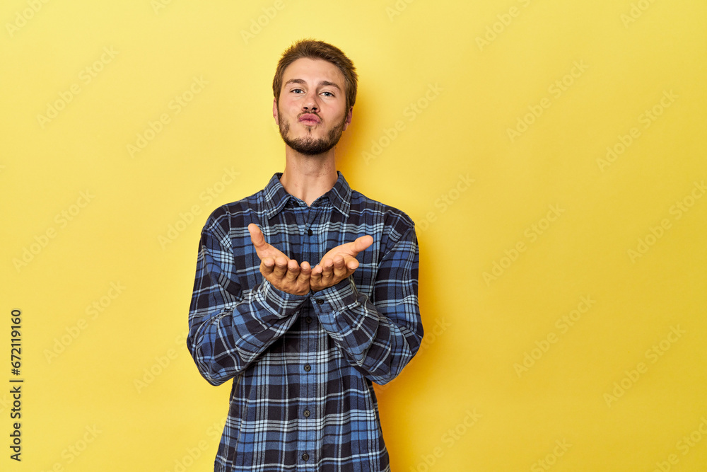Young Caucasian man on a yellow studio background folding lips and holding palms to send air kiss. Young Caucasian man on a yellow studio background folding lips and holding palms to send air kiss.