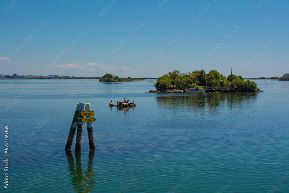 A sign post for Lignano and Aquileia in the Grado section of the Marano and Grado Lagoon in Friuli-Venezia Giulia, north east Italy. A fisherman's island and black cormorants on a rock can be seen in 