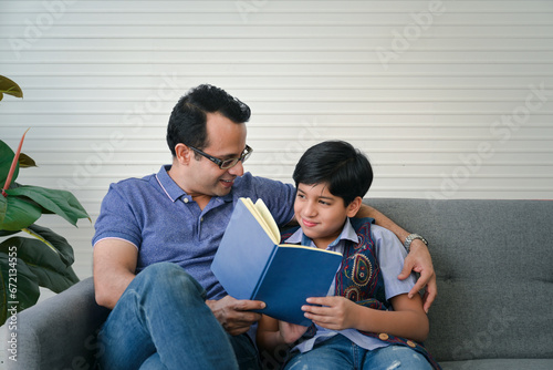 Happy indian father reading book with his son indoor, sitting on the coach and smiling at the living room.