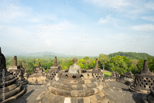 Obraz na płótnie (Selective focus) Stunning view of a Buddha Statue in the foreground and some bell shaped stupas in the background. Borobudur is a Mahayana Buddhist temple in Indonesia. w salonie