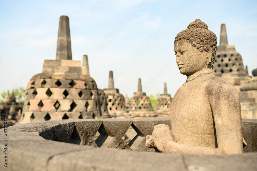 (Selective focus) Stunning view of a Buddha Statue in the foreground and some bell shaped stupas in the background. Borobudur is a Mahayana Buddhist temple in Indonesia. (Selective focus) Stunning view of a Buddha Statue in the foreground and some bell shaped stupas in the background. Borobudur is a Mahayana Buddhist temple in Indonesia.