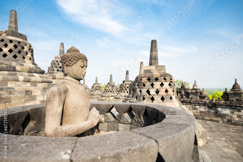Obraz na płótnie (Selective focus) Stunning view of a Buddha Statue in the foreground and some bell shaped stupas in the background. Borobudur is a Mahayana Buddhist temple in Indonesia. w salonie