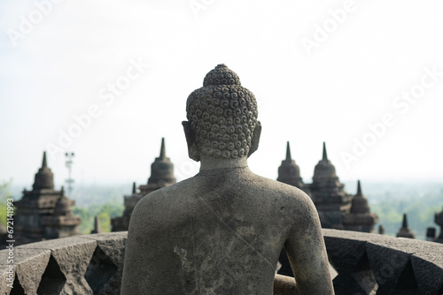 Obraz na płótnie (Selective focus) Stunning view of a Buddha Statue in the foreground and some bell shaped stupas in the background. Borobudur is a Mahayana Buddhist temple in Indonesia. w salonie