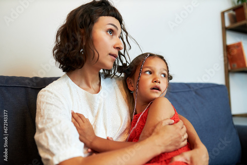Side view portrait of young mother supporting her child, hugging frightened little daughter on sofa scared of lightning and thunder outside looking aside with anxious facial expression photo