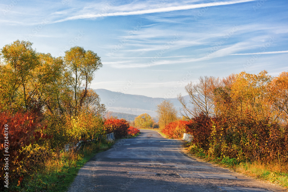 Fototapeta premium old cracked country road through rural valley in morning light. autumnal scenery with trees in fall colors on the roadside. mountains in the hazy distance