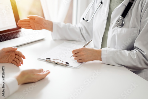 Doctor and patient sitting and discussing something near each other at the white desk in clinic. Female physician is pointing into laptop screen. Medicine concept