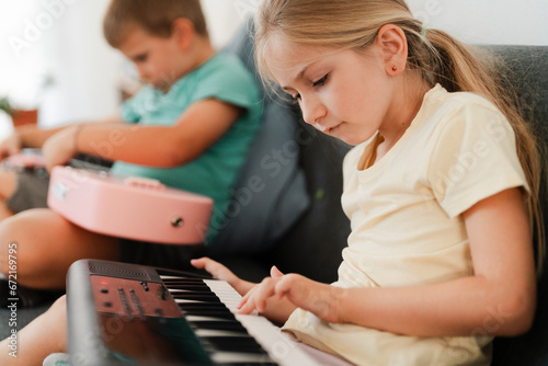 Side view of focused girl in casual clothes practicing piano while sitting near blurred boy playing guitar on sofa at home photo