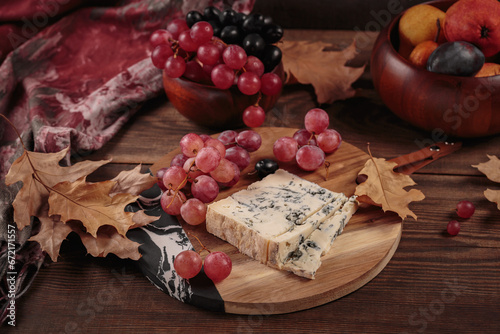 Cheese platter served with autumn fruits and dry leaves on dark wooden background. Appetizer of blue cheese on cutting board with grapes