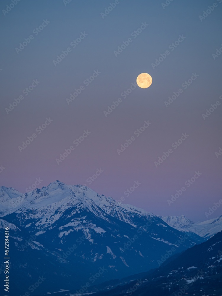 Snowy winter alpine landscape after sunset, pink purple sky with a full moon over a mountain peak