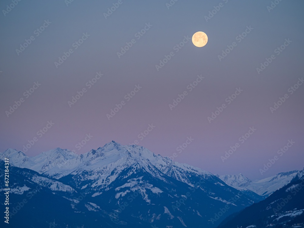 Snowy winter alpine landscape after sunset, pink purple sky with a full moon over a mountain peak Snowy winter alpine landscape after sunset, pink purple sky with a full moon over a mountain peak
