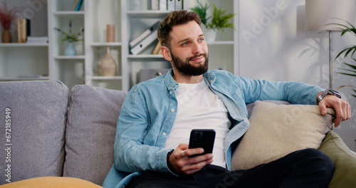 Happy handsome 30 old years man using smartphone in modern living room at home. Caucasian man resting on sofa while browsing the Internet and checking videos on social networks, indoors
