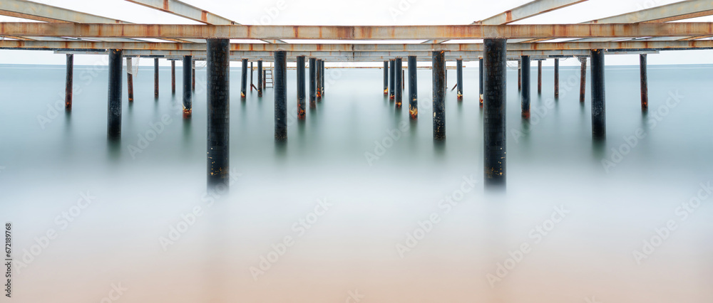 Fototapeta premium Fine art view of columns under metal pier at sunrise. Shot with long exposure to make the sea appear as fog