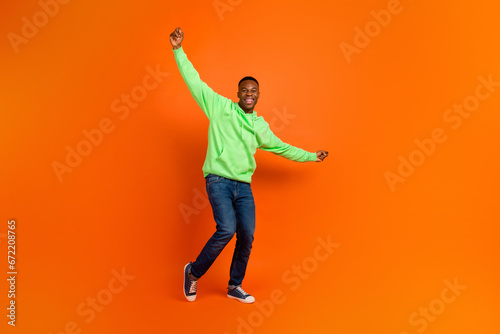 Full body photo of carefree good mood guy dressed green hoodie denim pants raising hands up win bet isolated on vivid orange background