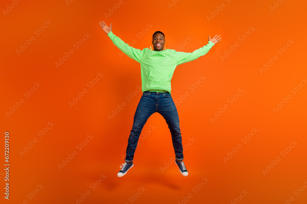 Full body photo of satisfied funky guy dressed green hoodie denim pants jumping demonstrate big sale isolated on vivid orange background