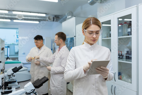 Mature confident and serious thinking woman in white medical coat working with tablet computer, team of scientists working in laboratory among microscope at workplace inside.