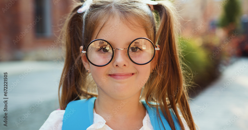 Close up. Portrait of lovely small school girl in glasses with backpack smiling posing on camera near campus. Female elementary school student holds textbooks standing outside school building. Close up. Portrait of lovely small school girl in glasses with backpack smiling posing on camera near campus. Female elementary school student holds textbooks standing outside school building.