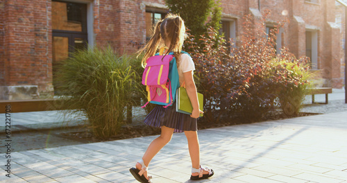 Little girl with backpack and textbook going hurrying to school. Attaractive schoolgirl with a backpack fun runs from school to home.