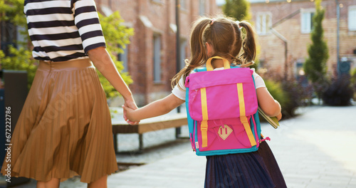 Back view young woman holds little girl in white dress by hand, go in city to school study, elementary class. Schoolgirl with backpack goes to school outdoor. Family back to school. Little girl goes