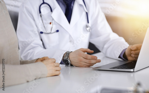 Unknown male doctor and patient woman discussing something while using laptop in a darkened clinic, glare of light on the background