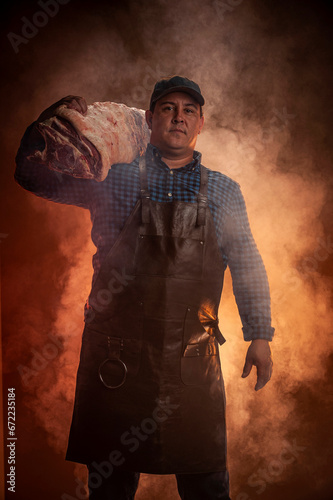 Butcher master. Middle aged hispanic man in apron holding meat with barbecue smoke background.