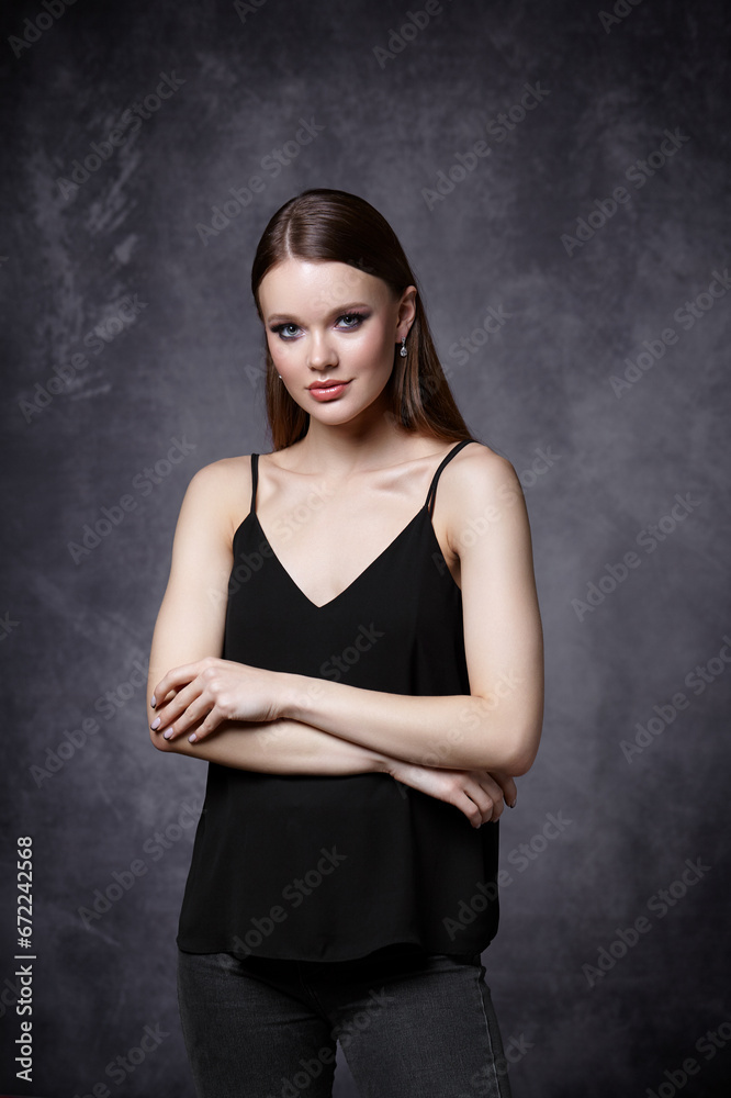 Portrait of female in black tank top with straps and jeans. Woman with earrings in the ear on gray background. Portrait of female in black tank top with straps and jeans. Woman with earrings in the ear on gray background.