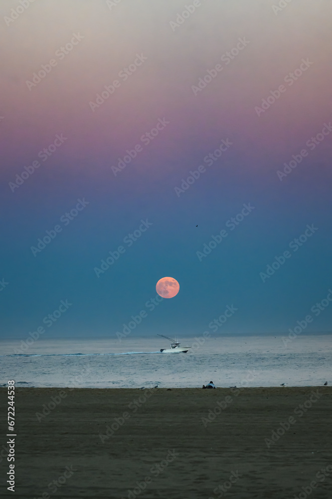 The full Harvest Moon is rising over the Atlantic Ocean as a photographer lies on the beach taking a photo of the moon and boat together. Ocean City MD vacation on the beach. Bordered The full Harvest Moon is rising over the Atlantic Ocean as a photographer lies on the beach taking a photo of the moon and boat together. Ocean City MD vacation on the beach. Bordered