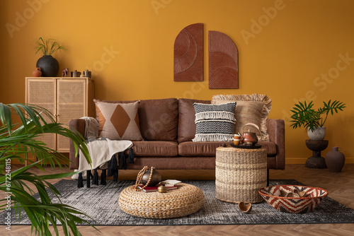 Warm and cozy composition of african living room interior with mock up poster frame, brown sofa, pouf, rattan sideboard, vase with leaves, pillows and personal accessories. Home decor. Template.