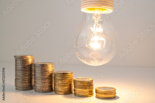 Lit lamp with stacks of coins of descending height. Reduction in energy tariffs.