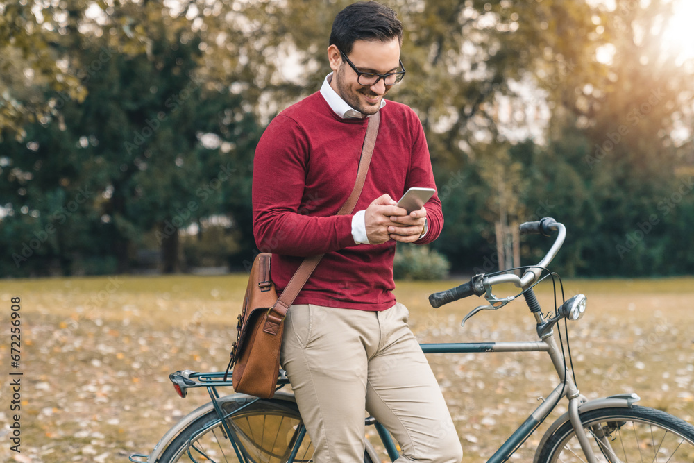 Smiling confident man using mobile phone while standing with his bicycle in public park on autumn day. Handsome businessman leaning against bicycle and texting.