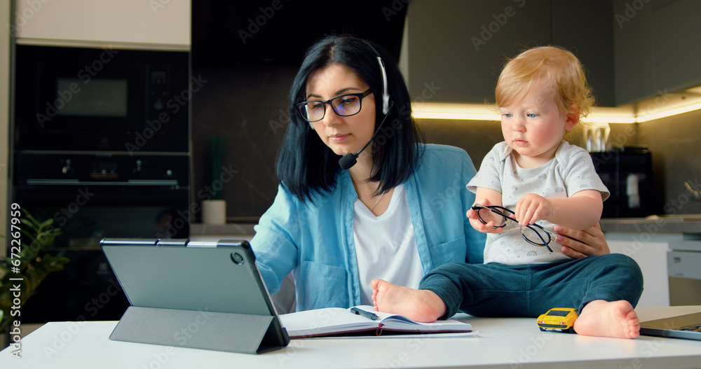 Woman in headphones using laptop and having online conference working from home. Mother and her little son are playing and working at the work table in the kitchen. Concept mom freelancer. Woman in headphones using laptop and having online conference working from home. Mother and her little son are playing and working at the work table in the kitchen. Concept mom freelancer.