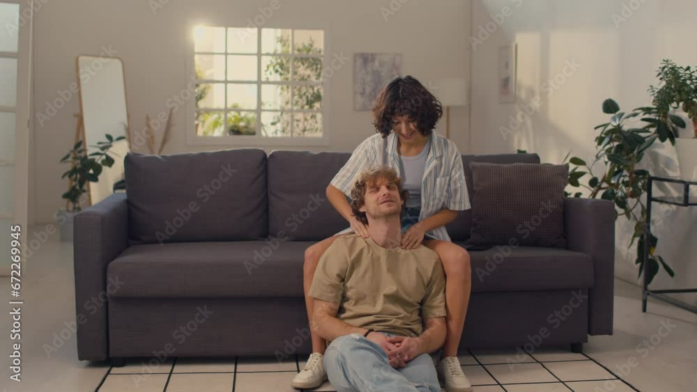 Young woman with black curly hair sitting on sofa giving shoulder massage to her boyfriend sitting on floor Young woman with black curly hair sitting on sofa giving shoulder massage to her boyfriend sitting on floor