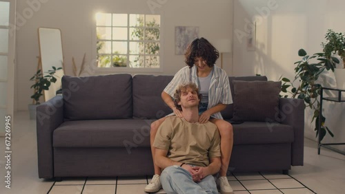 Young woman with black curly hair sitting on sofa giving shoulder massage to her boyfriend sitting on floor photo