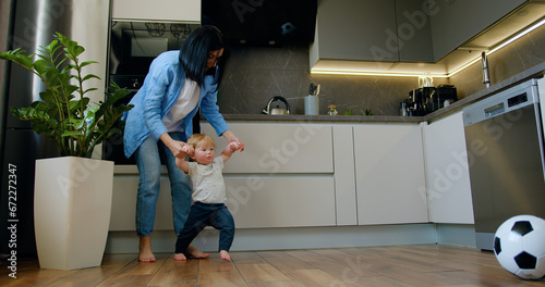 Mother helps the baby boy learn to walk. Child learns to take the first steps holding his mother's hands in modern kitchen at home. Happy family, mother support young toddler son.