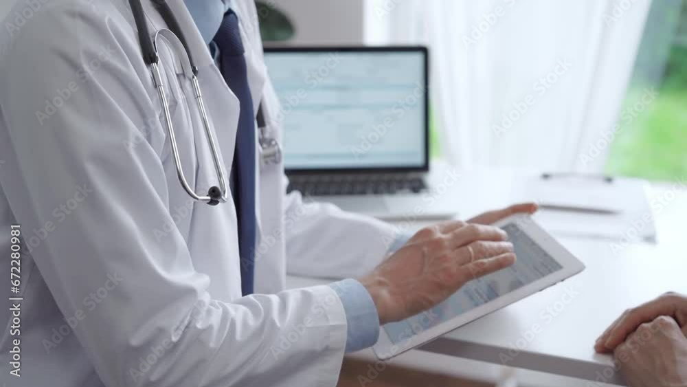 Doctor and patient are sitting at the table and talking in the clinic. Male pediatrician is using a tablet computer while discussing something with a middle-aged woman. Medicine, healthcare concepts Doctor and patient are sitting at the table and talking in the clinic. Male pediatrician is using a tablet computer while discussing something with a middle-aged woman. Medicine, healthcare concepts