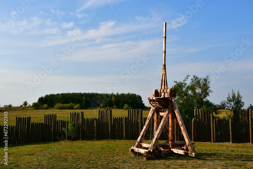 A close up on a ballista or a catapult made out of wooden logs, planks, and boards and serving as a medieval or viking artillery weapon seen on top of a hill covered with grass and trees in Poland