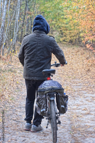 one male tourist in gray black clothes and a blue hood stands and holds a bicycle on a sand road in the autumn forest