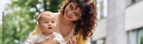 фотографія woman with wavy hair smiling near cute toddler daughter on city street, happy mo