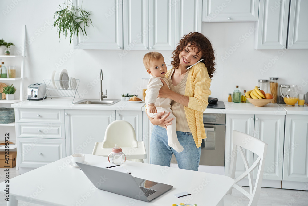 woman holding baby and talking on smartphone near laptop in modern kitchen, work life balance woman holding baby and talking on smartphone near laptop in modern kitchen, work life balance