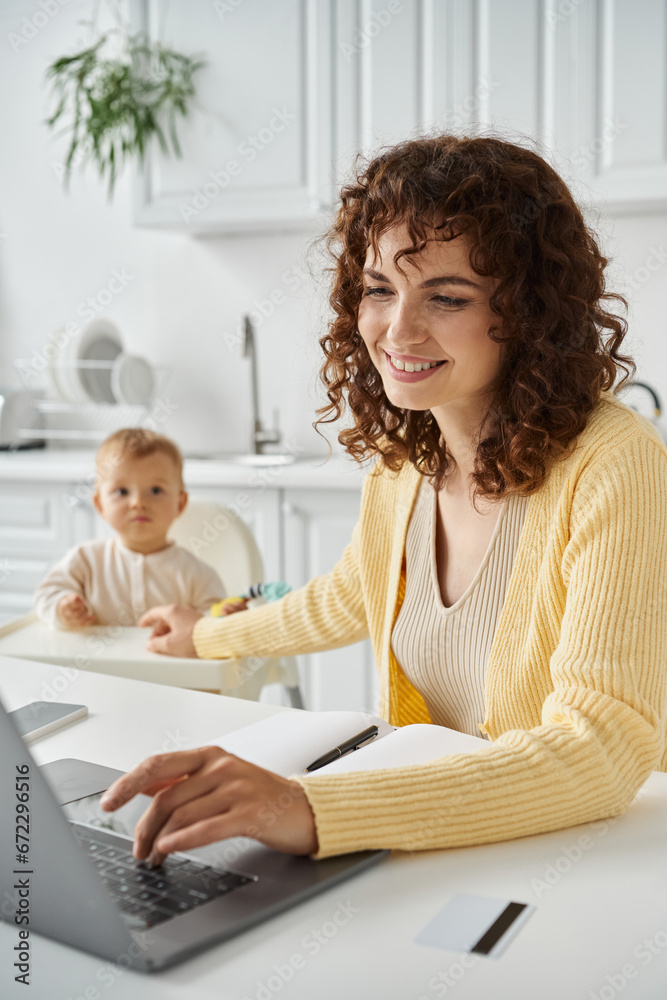 happy female freelancer working on laptop near little child in kitchen, work and family balance happy female freelancer working on laptop near little child in kitchen, work and family balance